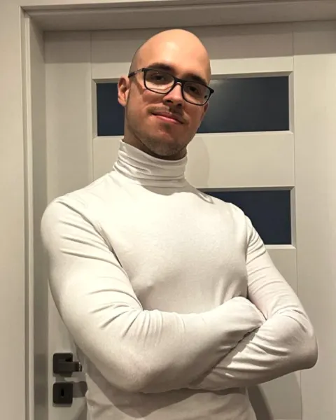 Krzysztof Piątek w białym golfie i okularach, z założonymi rękami.